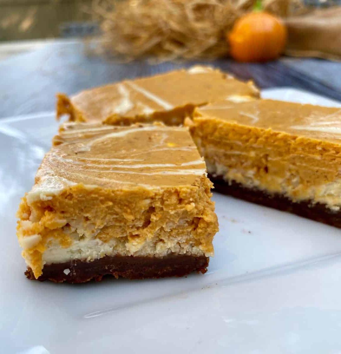 Three portions of pumpkin cheesecake bars on a plate with a small pumpkin and straw in the background.