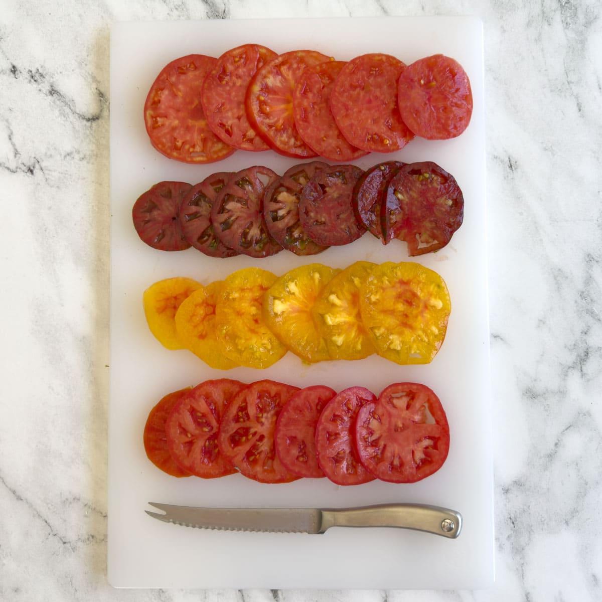 White cutting board on a marble counter with slices of colorful large tomatoes and a knife.