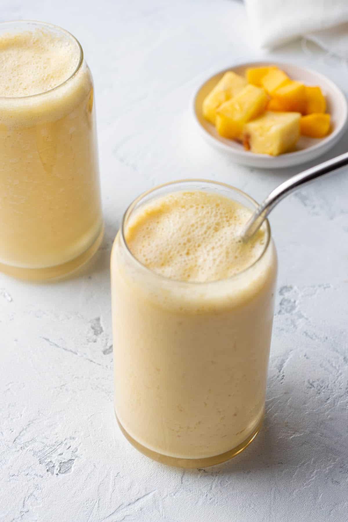 Two glasses with metal straws and a yellow smoothie with a plate of mangos in the back.