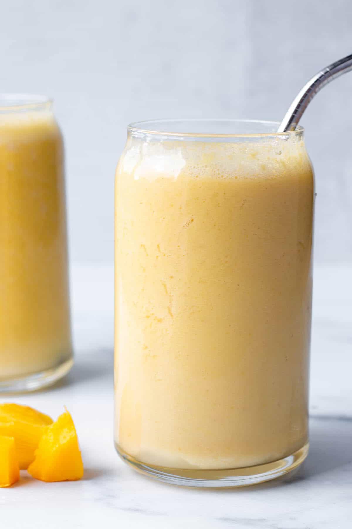 Two tall glasses with a mango smoothie, metal straw and fresh mango chunks.
