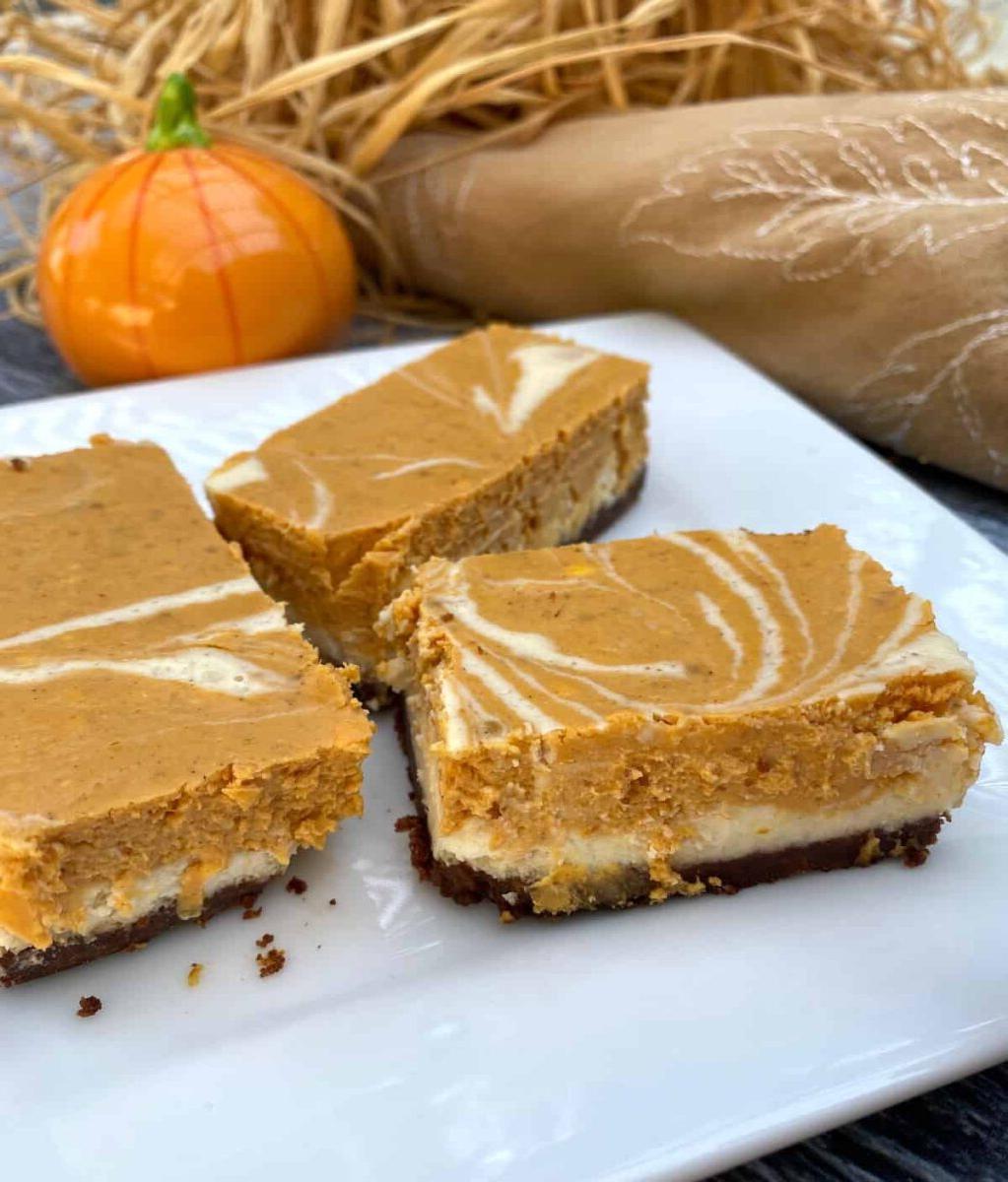 Three pumpkin cheesecake bars on a plate with straw and a miniature pumpkin ornament in the background.