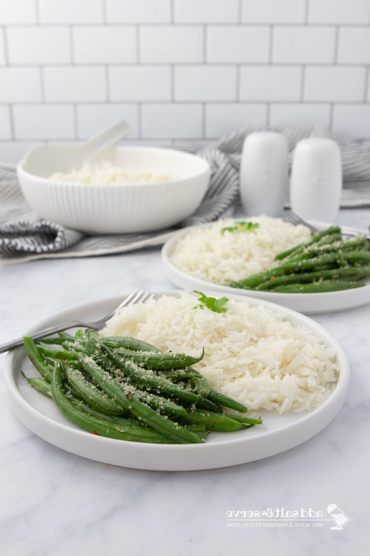 Seasoned green beans garnished with Parmesan cheese on plates with white rice with a serving bowl of rice in the background.